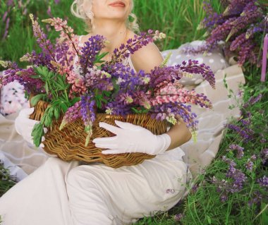 Beyaz uzun elbiseli gelin elinde bir sepet lupinle çimlerin üzerinde oturuyor.