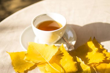 Beyaz bardakta çay ve sarı sonbahar yaprakları beyaz masada.
