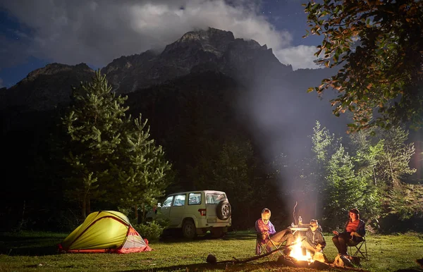 Família de três em um acampamento com fogueira à noite em terreno de montanha . — Fotografia de Stock