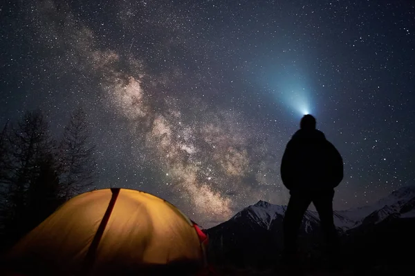 男子游客与手电筒站在他的帐篷附近, 晚上在一个充满星星的天空下. 图库图片