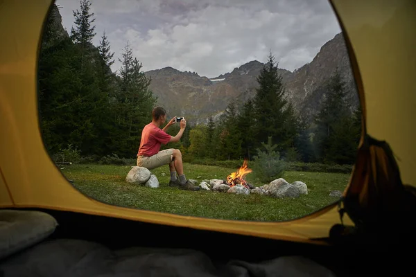 Vista de dentro da tenda turística. Homem caminhante sentado na fogueira, fazendo foto selfie pelo telefone móvel — Fotografia de Stock