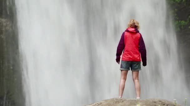 大きな滝の近くに赤いジャケットを着たハイキング女性 — ストック動画