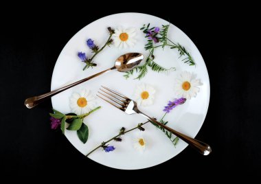 Çatal ve kaşıkla tabakta papatya ve yabani çiçekler. Siyah arka plan. Soyut ve gıda konsepti.