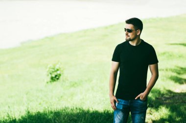 Sokaktaki gözlüklü adam. Genç bir adamın portresi. Yazın doğada yaşayan bir adam. Yürüyor, ayakta duruyor ve kahve içiyor. Siyah askılı ve kot pantolonlu yakışıklı bir adam. Ciddi bir adam başka yere bakar..