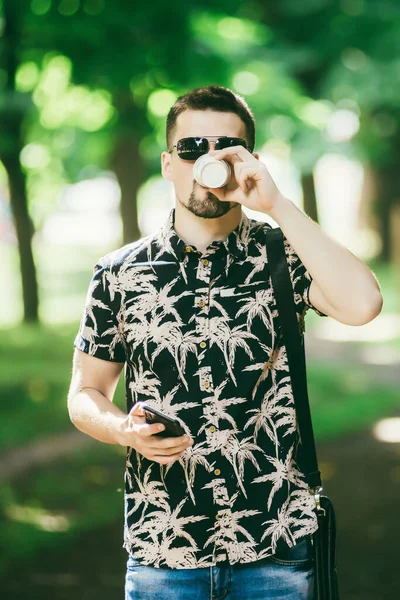 The guy with glasses on the street. Portrait of a young guy. Man on the nature in the summer. Walks or stands and drinks coffee. Handsome man in a black tank top and jeans. A serious man looks away.