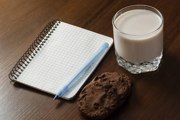 Flatley. A glass of milk, cookies, cinnamon and a notebook with a pen on a dark wooden table. White piece of paper with space for text. Diet and nutrition. Brown color. — Stock Photo, Image