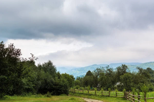 Köyün doğası. Ağaçlar, sisli arka planda dağlar. Yeşil ve mavi renkler. Çit ve yol kahverengi. Sakinleş ve dinlen. Bulutlar. Kasvetli gökyüzü