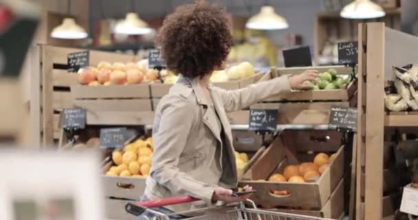 Afro-Amerikalı kadın gece yarısı market alışverişi yapıyor ve akıllı telefon kullanıyor. — Stok video