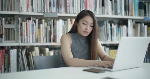 Asijská ženská pracuje v univerzitní knihovně na přenosném počítači a přijímá zprávu na telefonu — Stock video