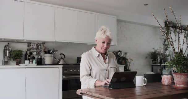 Ώριμη ενήλικη γυναίκα στην κουζίνα χρησιμοποιώντας ένα ψηφιακό tablet με πληκτρολόγιο — Αρχείο Βίντεο