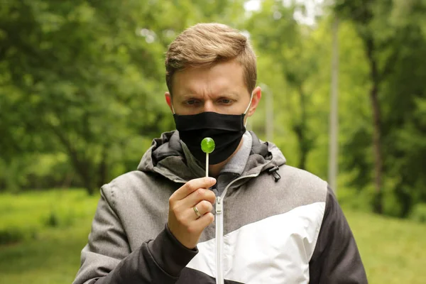 Masked man in the park. Quarantine problem. A man cannot eat a lollipop. Discomfort due to facial mask. Black mask guy