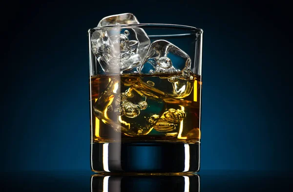 Viski eski moda bardakta, koyu mavi arka planda buz küpleri. Alkol içeceği, yansıma.
