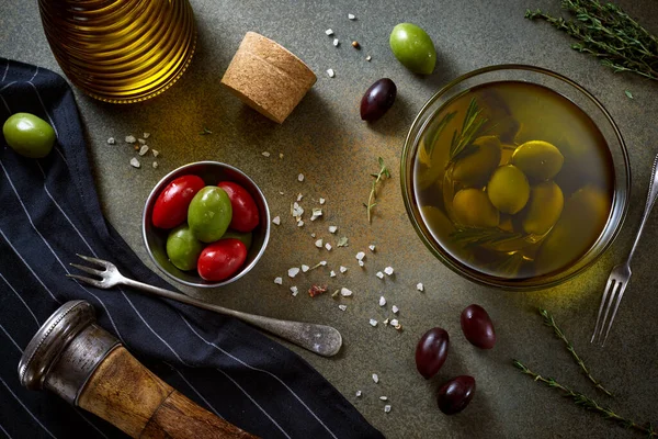 Kase içinde yeşil ve kırmızı büyük zeytinler. Manzara, yemek fotoğrafçılığı. Yeşil gride berbat bir sahne. Baharatlar, tuz, antika biber kutusu, yağ şişesi, çatal, biberiye ve kekik..