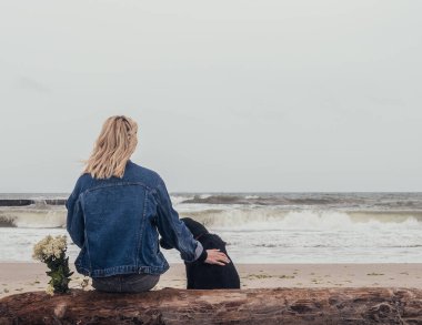 Genç bir kız ve siyah bir labrador bir kütüğe oturup denize bakıyorlar.