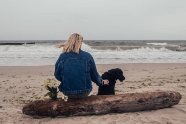 Genç bir kız ve siyah bir labrador bir kütüğe oturup denize bakıyorlar.