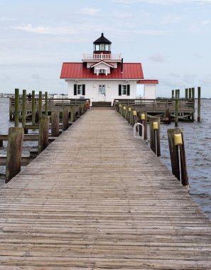İskelenin sonunda kırmızı çatılı ve beyaz kaplamalı küçük bir deniz feneri. Bir kaç hafif bulut, açık mavi gökyüzünde..