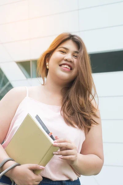 Portrait of Asian pretty smiley face fat woman pose and holding booklet front of building.