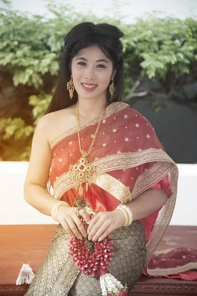 中央タイの古典的な伝統的なドレスに合わせて座っているポーズでかなりタイの女性が公園でスツールにガーランドを保持します — ストック写真