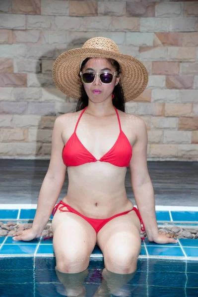 Mujer asiática bonita con bikini rojo sentada en el borde de la piscina con sombrero de paja y gafas de sol . — Foto de Stock