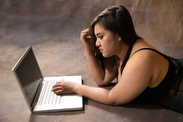 Asiática más tamaño sexy, gorda y con sobrepeso modelo de pelo largo femenino en vestidos negros en marrón estudio pose acostado uso portátil . —  Fotos de Stock