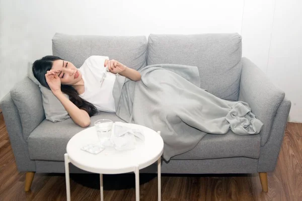 Όμορφη Ασιατική γυναίκα άρρωστο από το κρύο και χρησιμοποιείται με θερμόμετρο που βρίσκεται στον καναπέ στο δωμάτιο. — Φωτογραφία Αρχείου