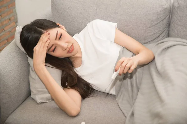 Pretty Asian woman sick from cold and used with thermometer lying on sofa in the room. — Stock Photo, Image