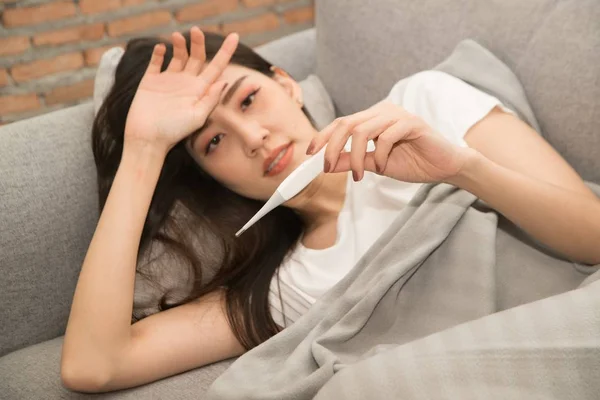 Szép ázsiai nő beteg hideg és használt hőmérő feküdt a kanapén a szobában. — Stock Fotó