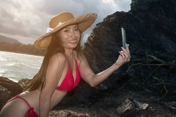 Asijská žena v plavkách v mobilním telefonu se staví a ona si bere s telefonem smartphone na pláži. — Stock fotografie