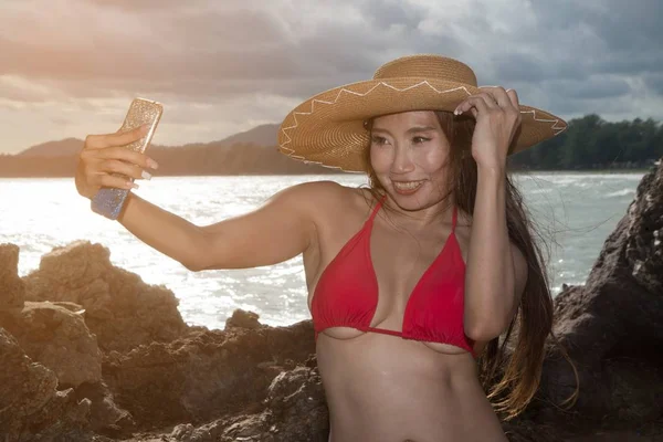 Una mujer asiática en un traje de baño de teléfono móvil se posa, se está tomando una selfie con un teléfono inteligente en la playa . — Foto de Stock