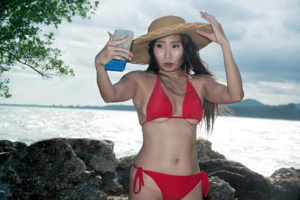 携帯電話の水着を着たアジア人女性が自分のポーズをとり、ビーチでスマートフォンでセルフィーを撮っている. — ストック写真