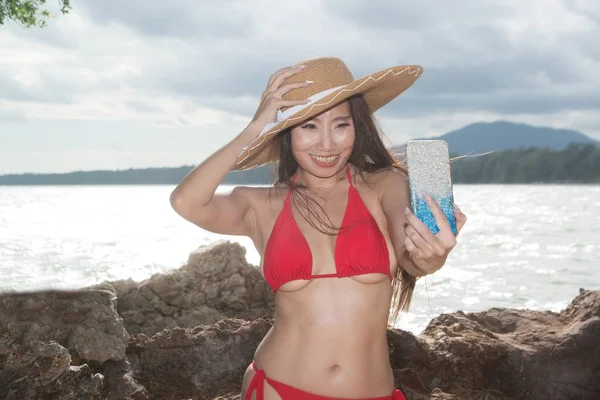 Азиатка в купальнике на мобильном телефоне позирует, делает селфи со смартфоном на пляже . — стоковое фото