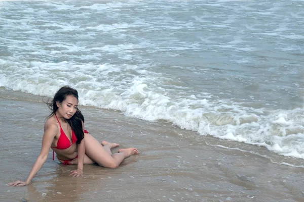 Una mujer asiática en un traje de baño rojo es pose sentada en una playa . — Foto de Stock