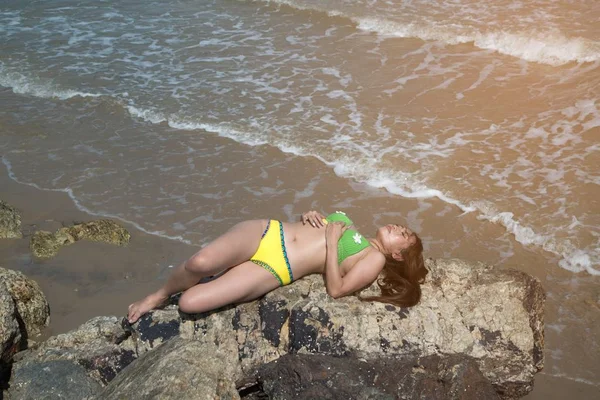一个穿着泳衣的亚洲女人躺在海滩上的一块石头上. — 图库照片