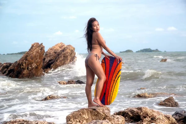 Mayo giymiş Asyalı bir kadın sörf tahtası yla ayakta duruyor ve sahilde bir kaya pozu vermek.. — Stok fotoğraf