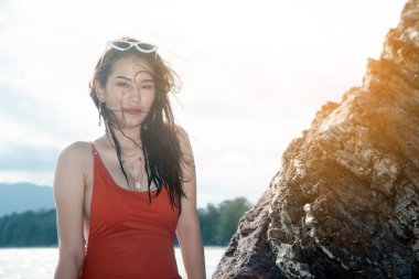 Kırmızı mayo yla Asyalı güzel kadın sahilde bir kayanın üzerinde oturuyor..