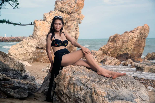 Szép ázsiai nő egy fekete fürdőruhát ül egy szikla a tengerparton. — Stock Fotó