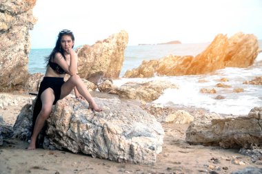 Siyah mayo pretty Asyalı kadın sahilde bir kaya üzerinde oturur. 