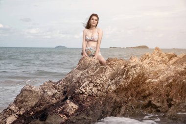 Ayakta bir mayo pretty Asyalı kadın ve plajda bir kaya üzerinde kumaş gösteriş. 