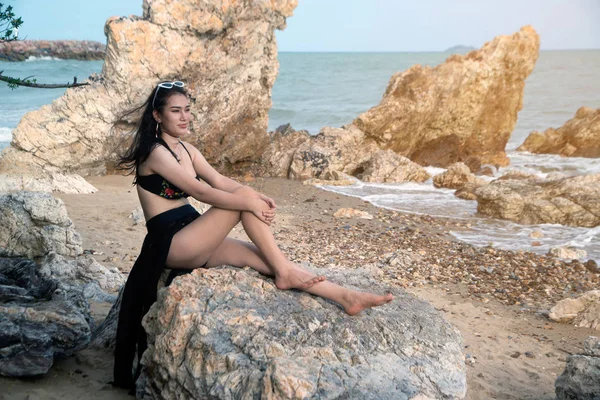Hezká asijská žena v černém plaveckém obleku sedí na skále na pláži. — Stock fotografie