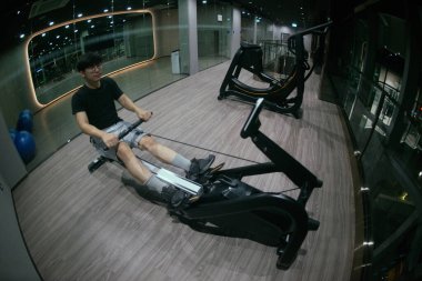 Formda genç bir erkeğin fitness stüdyosunda kürek çekme makinesiyle egzersiz yapması ön planda. Parlak modern spor salonu. Egzersiz yapan sağlıklı erkek kavramı.. 