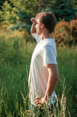 Genç adam uzun çimlerin üzerinde duruyor ve güneşe bakıyor.