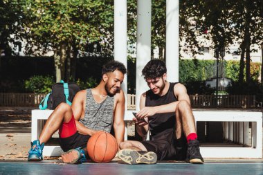 Genç bir adam, basketbol maçına ara verdiklerinde telefonunu başka bir delikanlıya gösteriyor.