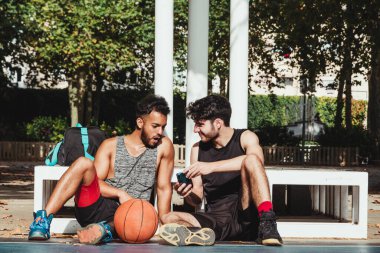 Genç bir adam, basketbol maçına ara verdiklerinde telefonunu başka bir delikanlıya gösteriyor.