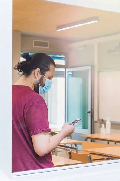 teacher with a face mask checks his schedule on his smartphone in an empty classroom