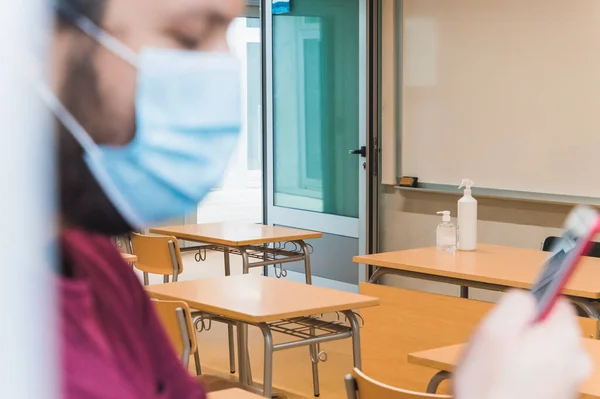 teacher with a face mask checks his schedule on his smartphone in an empty classroom