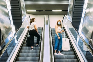 Yüz maskesi takan iki genç turist metro yürüyen merdivenlerini kullanıyor.