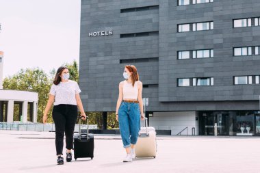 Maskeli iki genç turist Covid-19 nedeniyle güvenli bir mesafede kalarak otelden ayrılıyor.