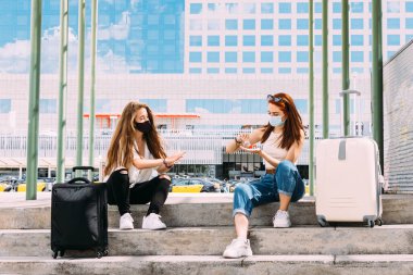 Yüz maskesi takan iki genç turist güvenli bir mesafede oturuyorlar çünkü covid-19 ellerini temizlemek için anti-bakteri jeli kullanıyorlar.