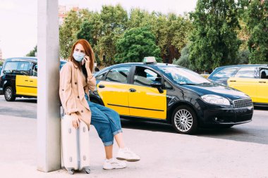 Bagajında oturan genç bir kadın, taksi beklerken telefonla konuşuyor.