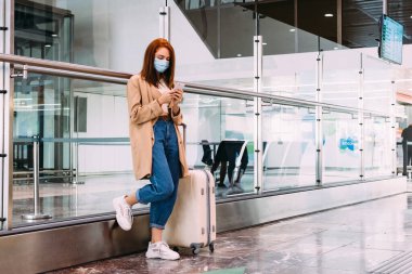 Maskeli kadın tren istasyonunda akıllı telefonunu kullanarak mesaj gönderiyor.